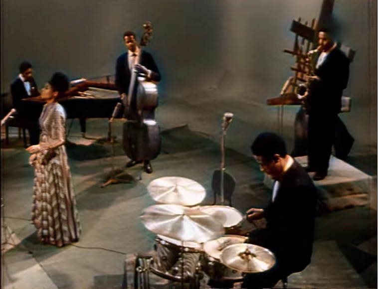View of the Max Roach group from stage-left, downward facing camera angle of full group including drums, bass, piano, tenor sax and Abbey Lincoln on vocal